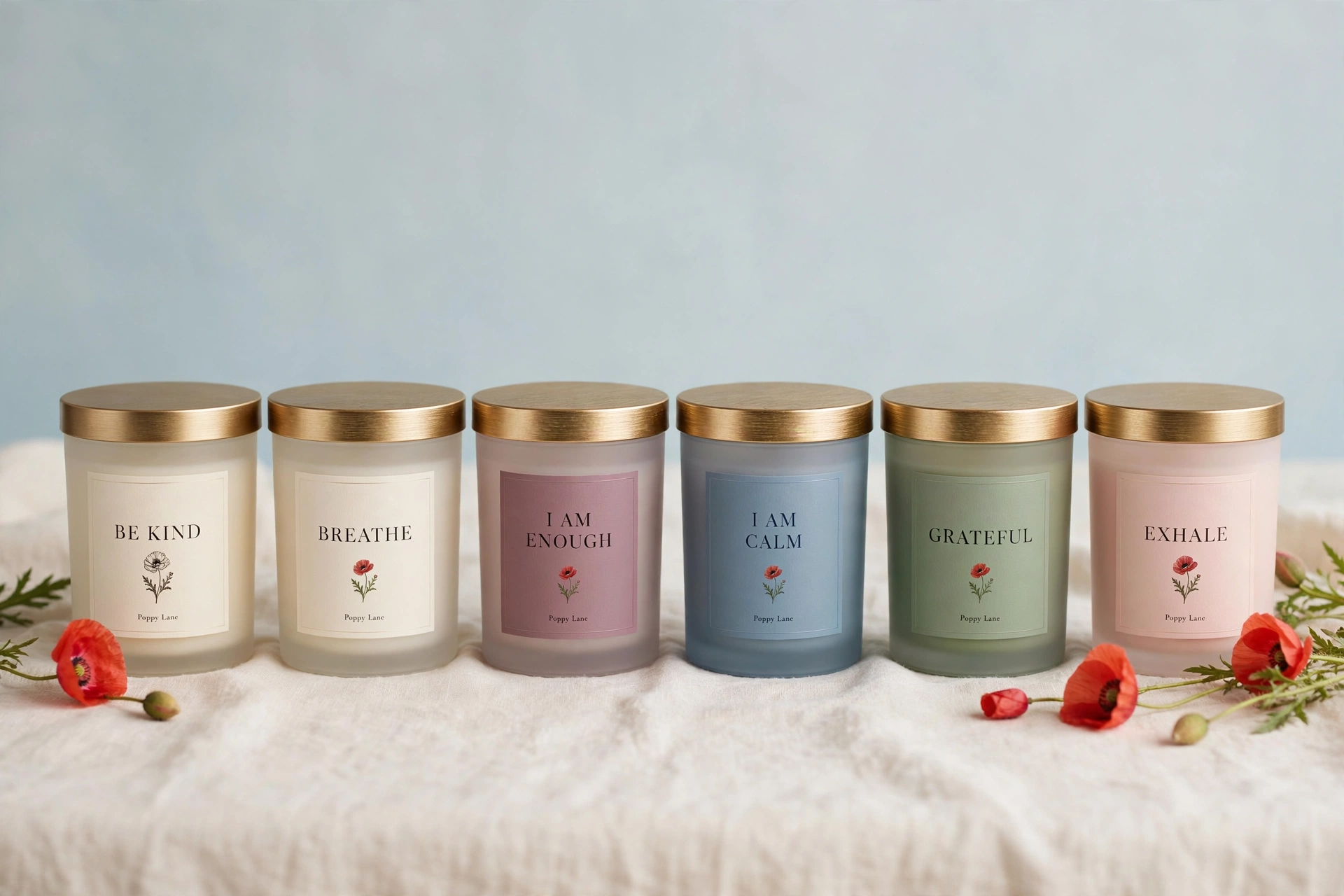 Rendering of the six Poppy Lane word candles in frosted glass jars with gold lids on cream linen: two cream labels reading Be Kind and Breathe, and four colored labels, I Am Enough in mauve, I Am Calm in dusty blue, Grateful in sage, and Exhale in blush, each with a small poppy and the words Poppy Lane