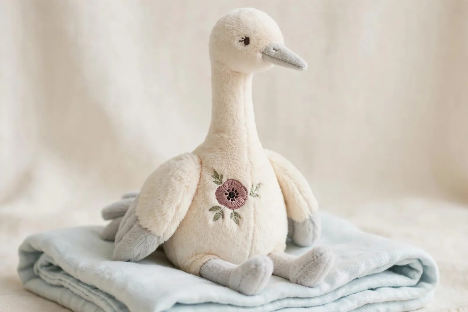Soft cream plush crane toy with a long neck, gray wing tips and a dusty-pink embroidered poppy on its chest, sitting on a folded pale-blue blanket