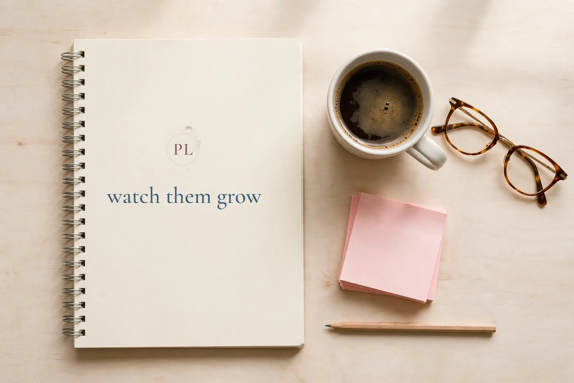 The Poppy Lane teacher planner lying closed on a desk: a cream spiral-bound cover with the PL monogram above the words watch them grow, beside a mug of coffee, reading glasses, and pink sticky notes